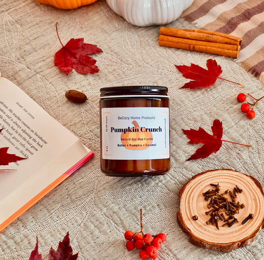 A jar candle labeled 'Pumpkin Crunch' by BeCozy Home Products is styled on a textured beige blanket with autumn props including red maple leaves, cinnamon sticks, a book, a wood slice with cloves, and small red berries.