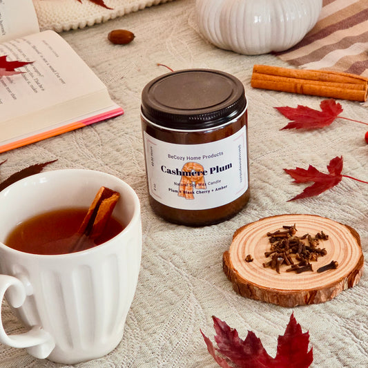A jar of BeCozy Home Products Cashmere Plum Candle sits on a textured beige blanket surrounded by cozy autumn props, including a white mug of tea with a cinnamon stick, scattered red leaves, cinnamon sticks, and a wood slice.