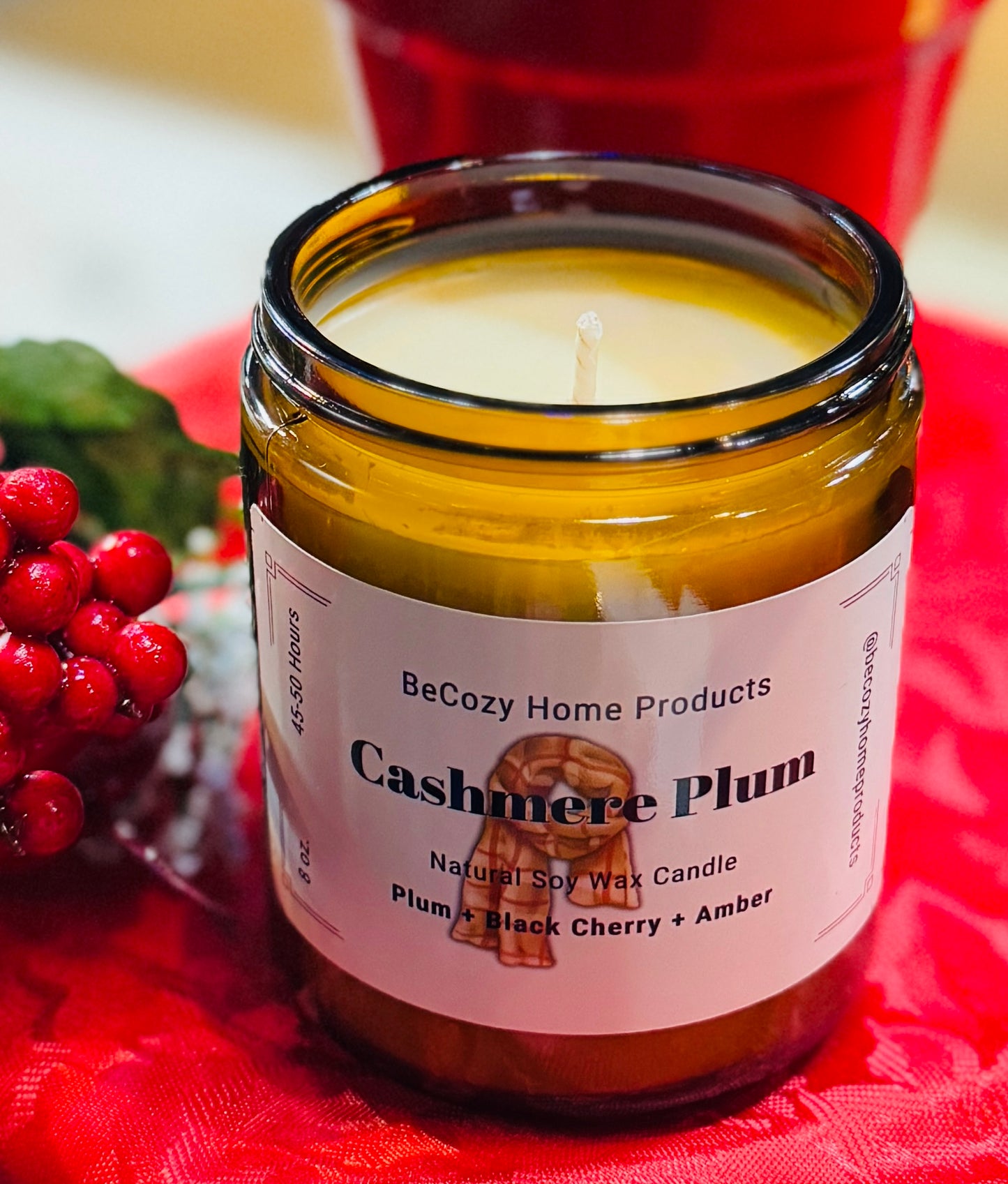 A soy wax candle with a label reading 'Cashmere Plum' placed on a red textured surface, with decorative branches and a red berry wreath in the background.