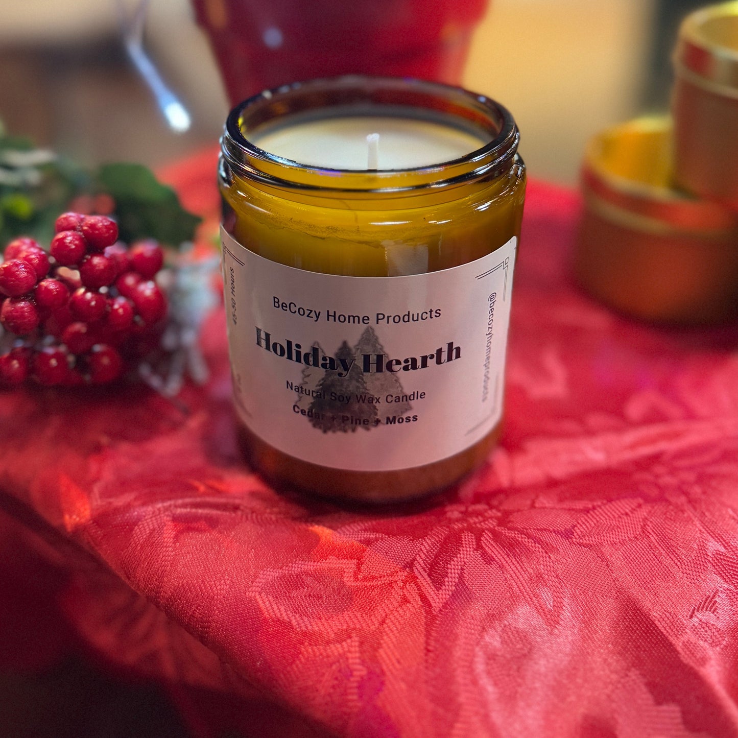 A jar of Holiday Hearth soy wax candle by Be Cozy Home Products, placed on a red textured surface with Christmas decorations around it.