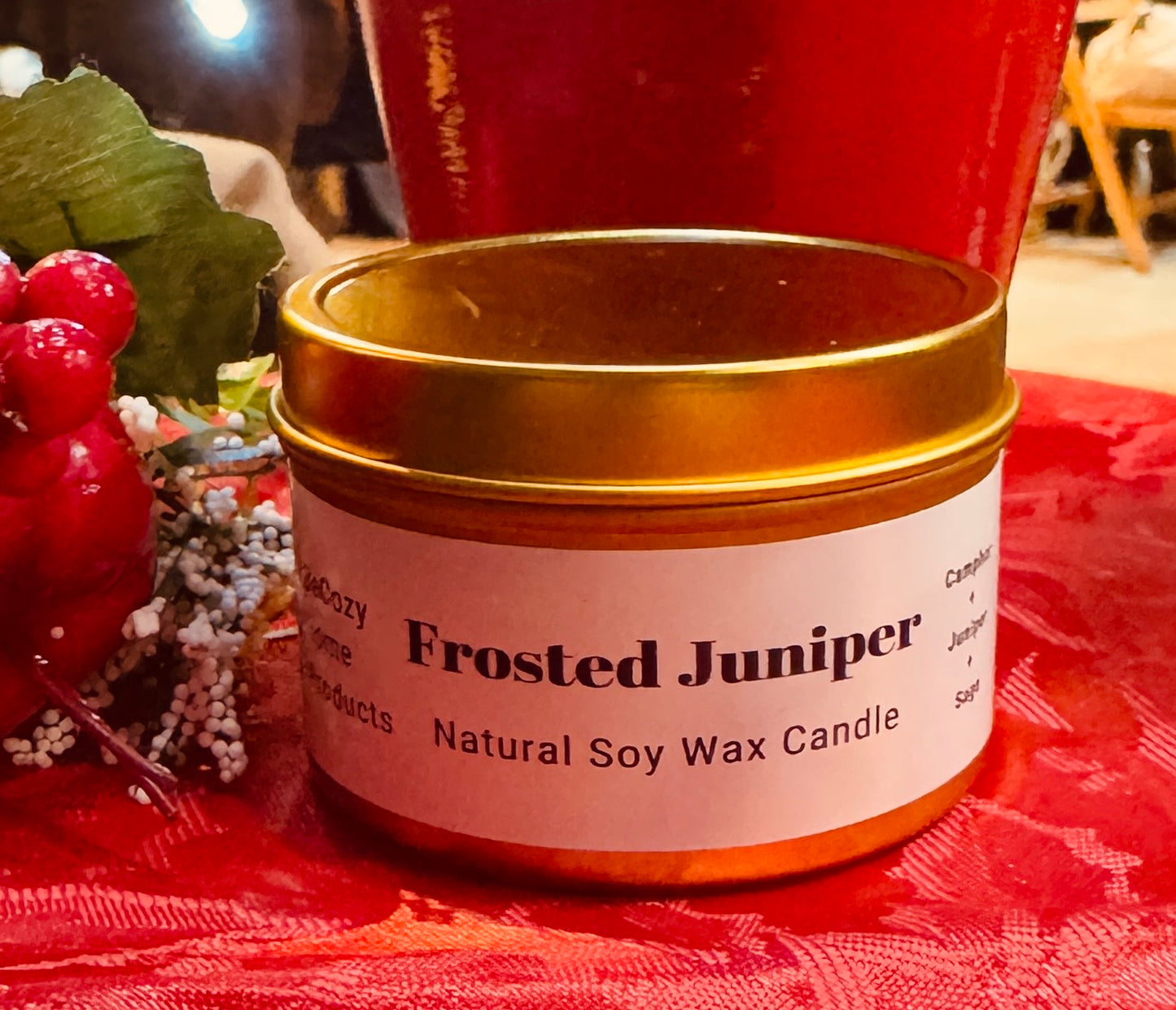 A soy wax candle with a label reading 'Frosted Juniper' placed on a red textured surface, with decorative branches and a red berry wreath in the background.