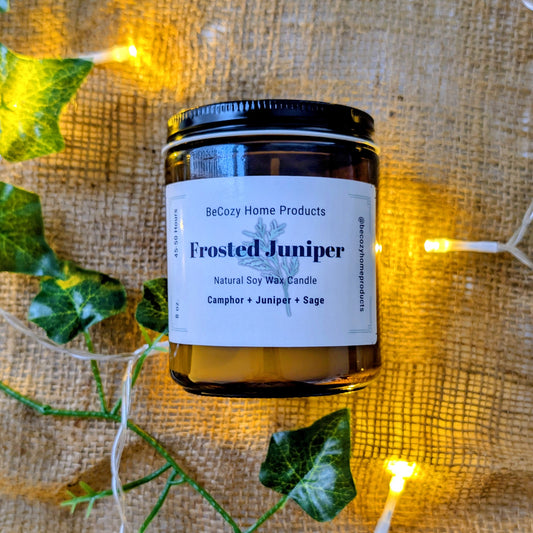 Soy Wax Candle labeled 'Frosted Juniper' by BeCozy Home Products on a textured fabric background with greenery and lights.