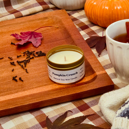 A small candle tin labeled 'Pumpkin Crunch' sits on a wooden tray in an autumn-themed setting with pumpkins, leaves, a mug, and a cozy plaid blanket.