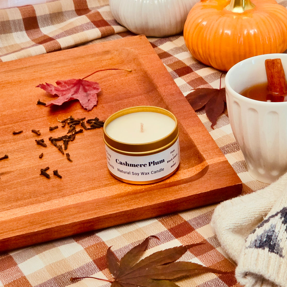 A small candle tin labeled 'Cashmere Plum' sits on a wooden tray beside a mug of tea, autumn leaves, cloves, and pumpkins on a plaid blanket.
