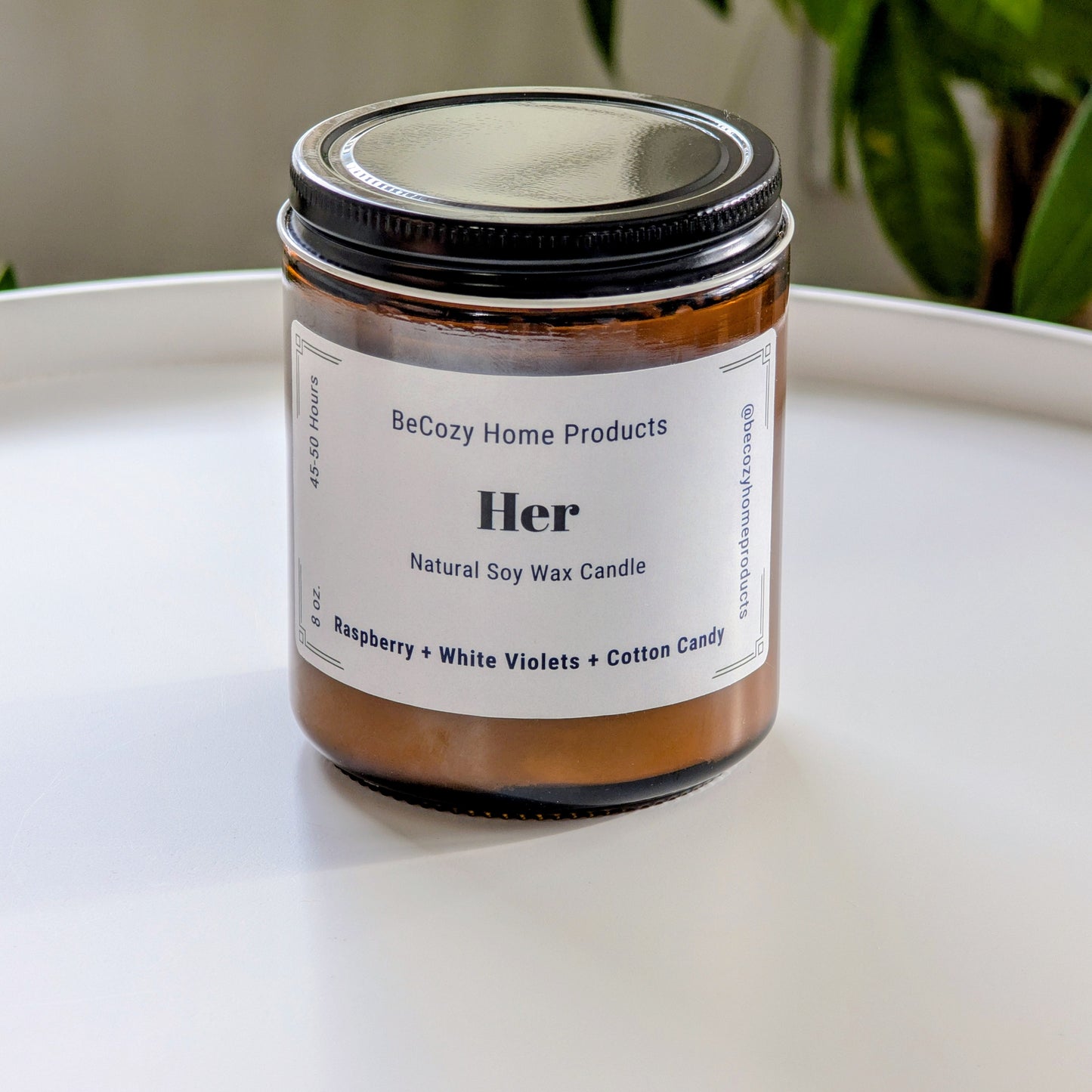 Soy Wax Candle jar labeled 'Her' by BeCozy Home Products on a white surface with a plant in the background.