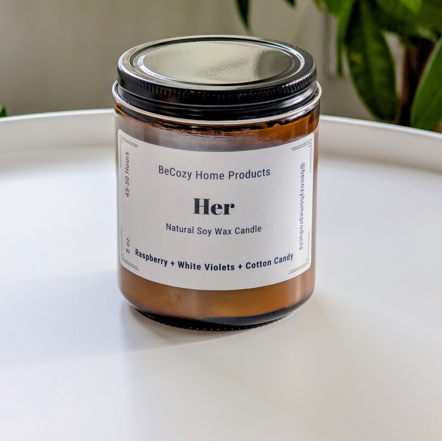 Soy Wax Candle jar labeled 'Her' by BeCozy Home Products on a white surface with a plant in the background.
