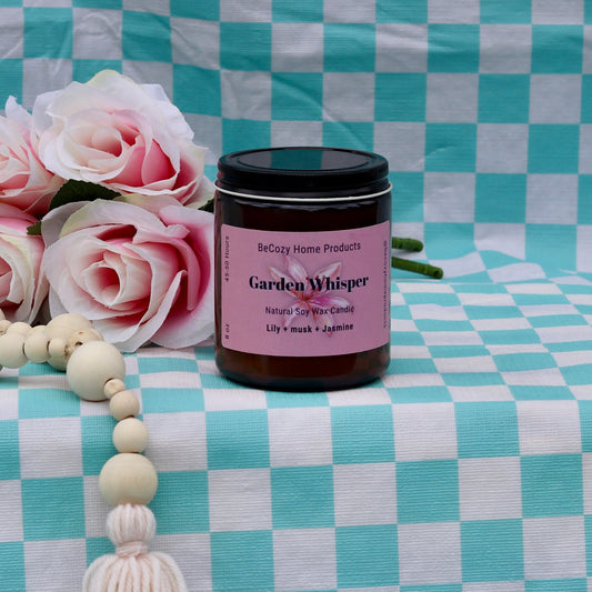 A jar of Garden Whisper - Soy Wax Candle with a label showing the product name and the scents of jasmine and lily. The jar is on a checkered fabric background.
