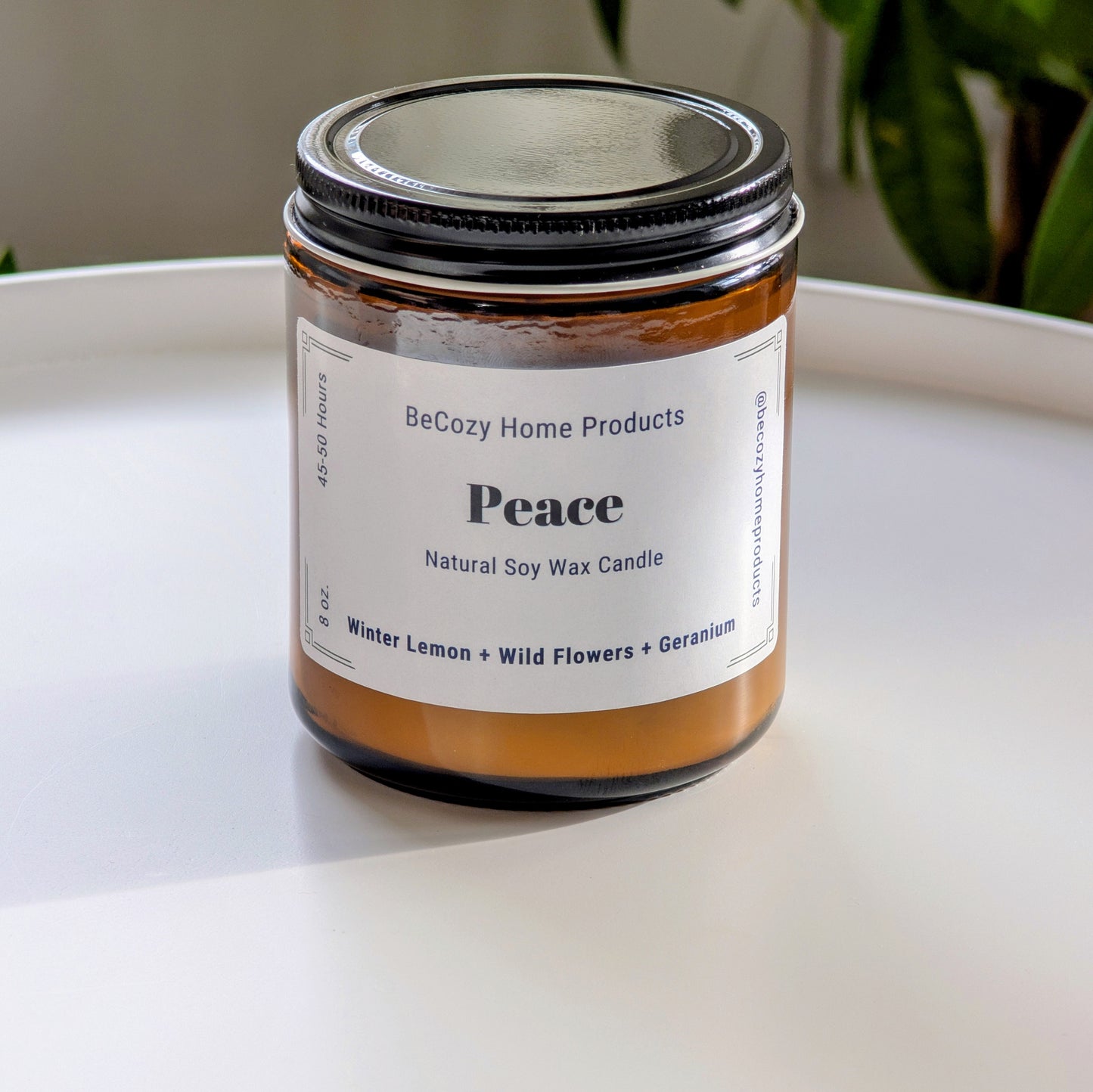 Soy Wax Candle jar labeled 'Peace' by BeCozy Home Products on a white surface with a plant in the background.