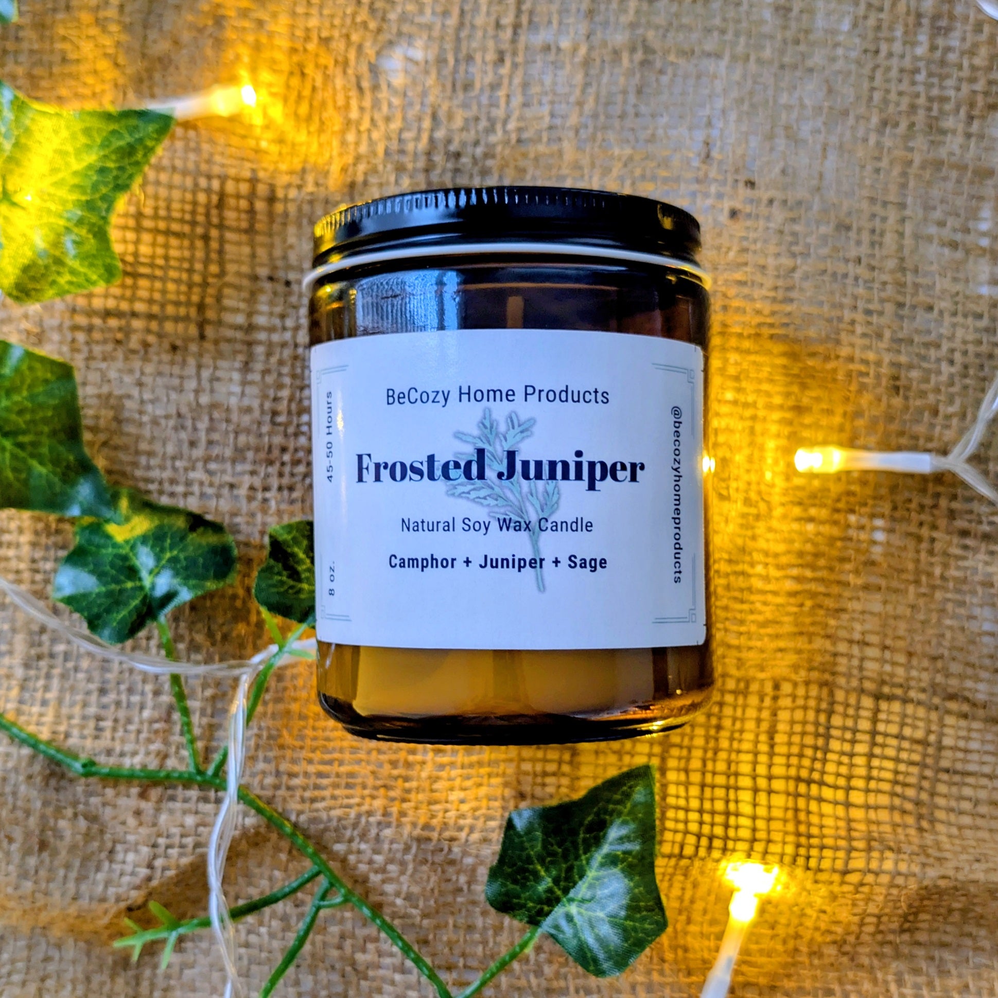 Soy Wax Candle labeled 'Frosted Juniper' by BeCozy Home Products on a textured fabric background with greenery and lights.